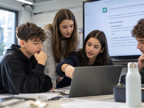 Vier Jugendliche arbeiten gemeinsam an einem Laptop. Im Hintergrund ist eine KI-Anwendung auf einem Bildschirm zu sehen.